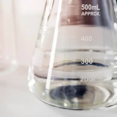 A close-up of a clear liquid being poured into a laboratory glass beaker or flask, representing industrial hydrosol chemical production.